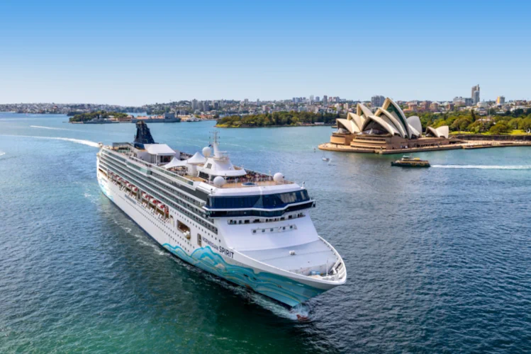 Bild mit Opernhaus: Ein großes Kreuzfahrtschiff mit bunter Rumpfbemalung fährt im Hafen von Sydney am berühmten Opernhaus vorbei.