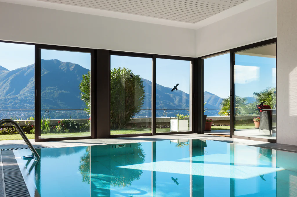 Ein Hallenbad mit klarem, blauem Wasser im Vordergrund, das die Fenster spiegelt. Große Glasschiebetüren bieten einen weiten Ausblick auf einen grünen Garten, Palmen und eine bergige Landschaft unter blauem Himmel.